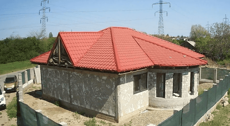 Clădire rezidențial în construcție cu acoperiș din țiglă roșie, deschidere spre curte și panoramă rurală.