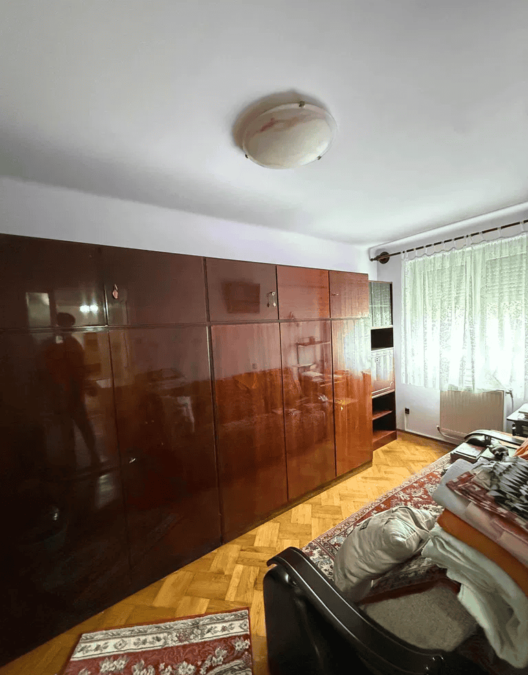 Dormitor cu mobilier din lemn lucios și perdele albe, lumină naturală, decor confortabil.