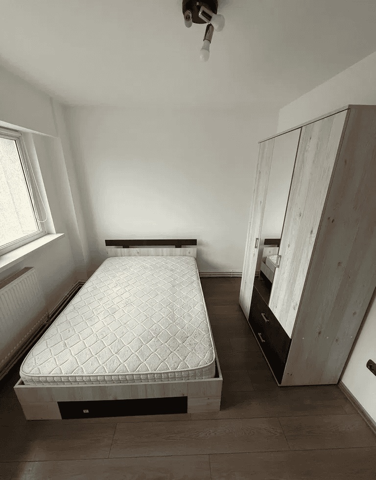 Dormitor cu pat și dulap spațios, lumină naturală, mobilier alb modern.